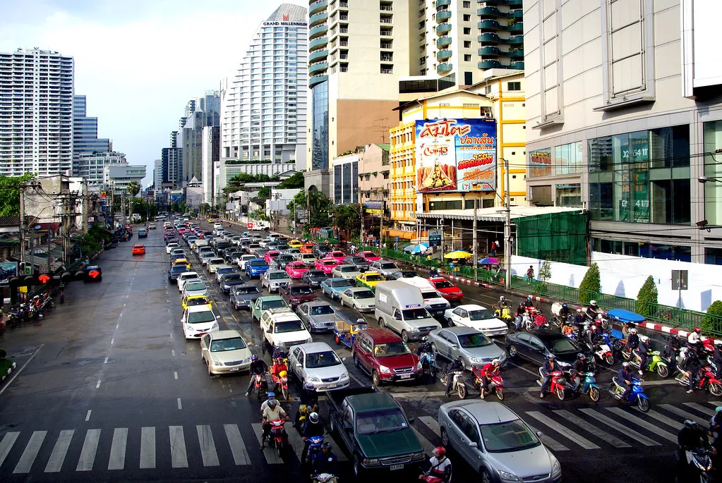 Cars, taxis and motorcycles queued at a crossing on a wide Bangkok road lined with tower blocks