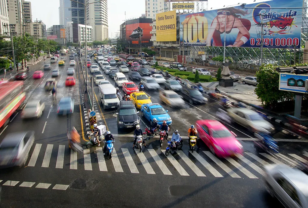 Busy Bangkok traffic seen from above, with cars, a yellow taxi and motorcycles crossing a junction